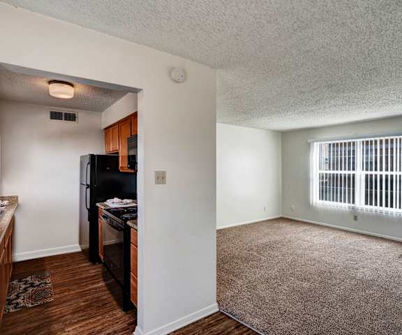 A view of the empty carpeted living room with a window and the kitchen with black appliances and wooden large cabinets at Golden Crest. 