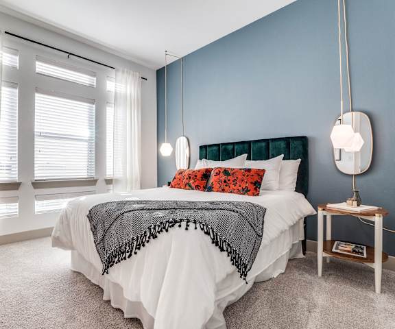A spacious bedroom with a queen-size bed with a white bedspread, a dresser, wall art, a light blue accent wall, a wicker chair, decorations, window, and nightstands with lamps at the Crosby at the Brickyard Apartment Homes. 