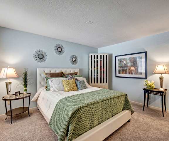 A spacious light blue bedroom with a queen-size bed with a green and white bedspread, a dresser, wall art, lamps, decorations, window, and nightstands at River's Cove Apartment Homes. 