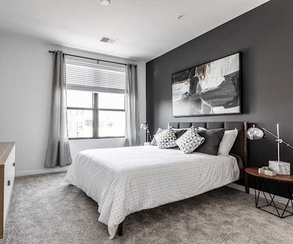 A spacious bedroom with a queen-size bed with a white bedspread, a dresser, wall art, lamps, decorations, window, a black accent wall, and nightstands at Vim + Vigor Lofts. 