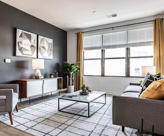 The spacious, modern, gray living room with a couch, throw pillows, large windows, a ceiling fan, wall art, plush chairs, coffee table, decorations, a TV, and large window with yellow curtains at Vim +Vigor Apartment Homes.