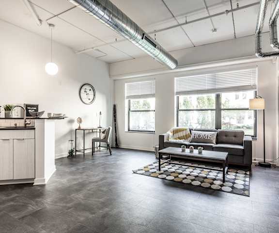 The spacious, gray, industrial living room with a couch, plush chairs, coffee table, overhead lighting with a ceiling fan, decorations, a TV, and view of the dining room with a table, chairs, and place settings for four, and the kitchen at Vim + Vigor Apartment Homes. 