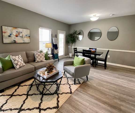 The living room with a couch, plush chairs, coffee table, decorations, a TV, windows, and view of the dining room at Harbin Pointe Apartment Homes.
