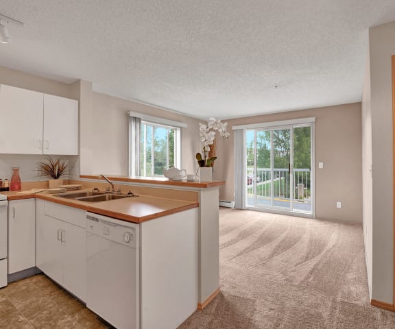 A view of the empty carpeted living room with a window and the kitchen with white appliances and large wooden cabinets at Manning Crossing Apartment Homes. 