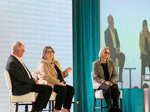 Three people are sitting on stage in front of a screen with another person on it.