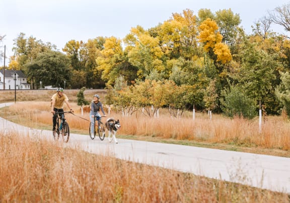 A man and a woman riding bikes down a road with a dog at Riverhouse Apartments, Fargo, ND 58102