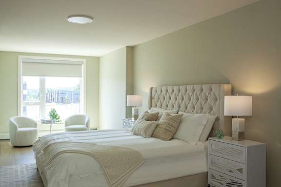 A bedroom with a large bed, a white chair, and a window with a view of the city.