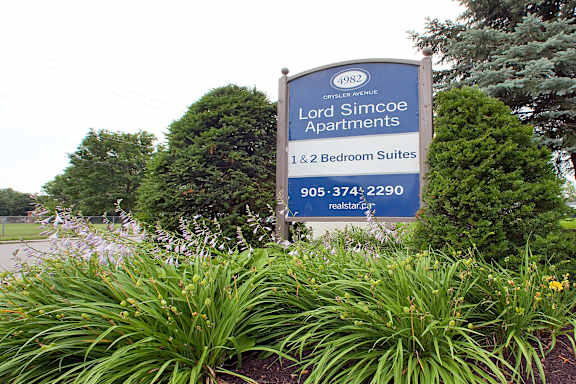 A sign for Lord Simcoe Apartments stands in front of some trees and plants.