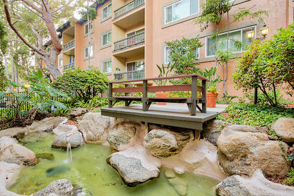a bench sits on a bridge over a stream in front of an apartment building