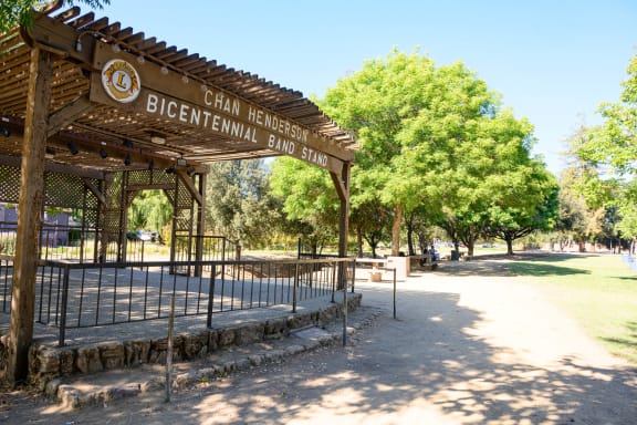 A wooden shelter with a sign that reads "Bicentennial Band Stand" is in the foreground of a sunny park. at Pleasanton Place, Pleasanton, CA
