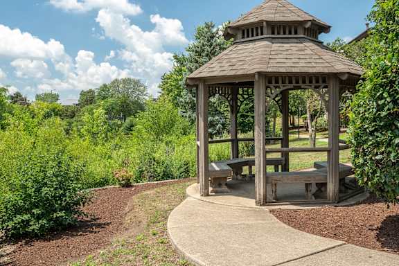 A wooden gazebo with a brown roof is surrounded by greenery and has a circular concrete walkway around it.