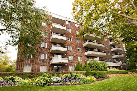 A large brick building with balconies and windows is surrounded by a well-kept garden with a variety of flowers.
