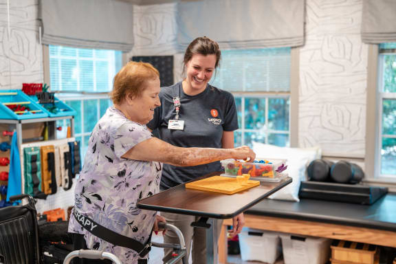 an older woman painting a picture at a table with other people painting on canvases