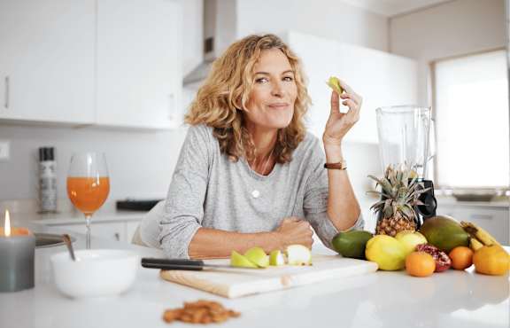 A woman is sitting at a kitchen counter with a bowl, a glass of orange juice, and various fruits and vegetables.