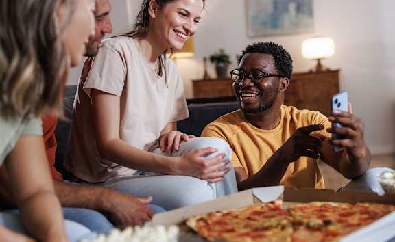 A group of people are sitting on a couch, eating pizza and playing a card game.