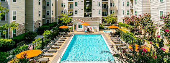 A large swimming pool surrounded by lounge chairs and umbrellas in front of apartment buildings.