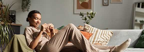 A woman is sitting on a couch with a green blanket, holding a book and a cup of coffee.