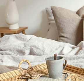A grey mug with a wooden handle sits on a woven tray with a white lamp and pillows in the background.