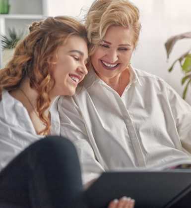 Two women are sitting on a couch, one is reading a book and the other is laughing.
