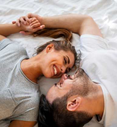 A couple is lying on a bed, holding hands and smiling at each other.