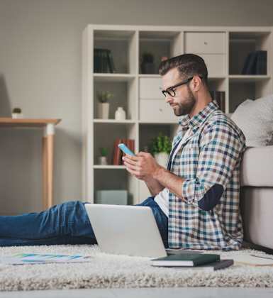 A man is sitting on the floor with his feet up, using a laptop and holding a smartphone.