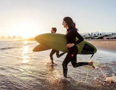 A person in a black wetsuit is running on the beach with a green surfboard and a yellow leash.