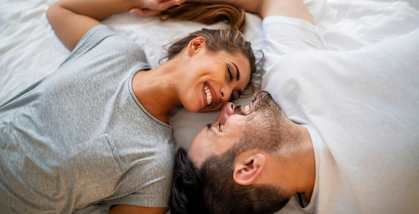 A couple is lying on a bed, holding hands and smiling at each other.
