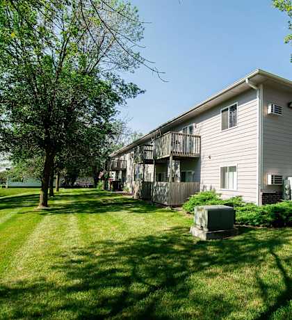 A house with a grey siding and a balcony is surrounded by green grass and trees at Peach Tree Apartments, Iowa, 50021
