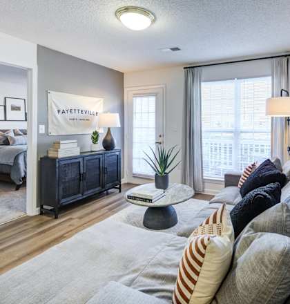 A living room with a grey sofa and a bedroom with a bed and a nightstand in the background.
