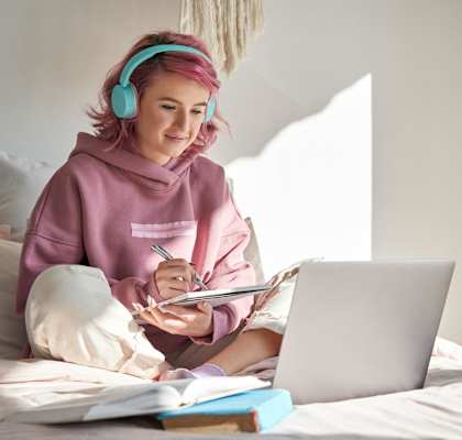 A person with pink hair is wearing headphones and writing in a notebook while using a laptop. at Creekview Homes, Chico, CA, 95928