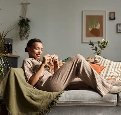 A woman is sitting on a couch with a green blanket, holding a book and a cup of coffee.