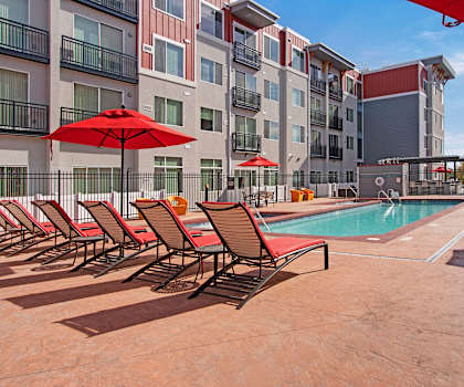 The large, turquoise outdoor pool with red pool chairs, red umbrellas, tables, and chairs with the apartments in the background at the Ritz Classic Apartment Homes. 