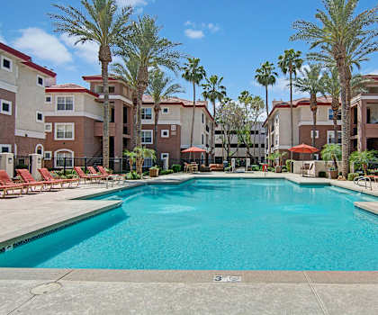 The turquoise outdoor pool with palm trees, orange pool chairs, tables, and chairs with the apartments in the background at Mozaic at Steele Park Apartments. 