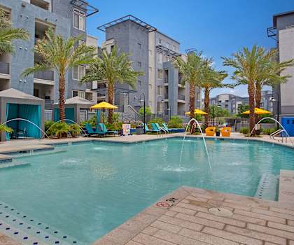 The large, turquoise outdoor pool surrounded by palm trees, pool chairs, tables, cabanas, and chairs with the apartments in the background at Rio Paradiso Apartment Homes. 