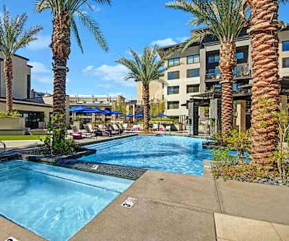 The large, turquoise, resort-style outdoor pool surrounded by palm trees, blue pool chairs, tables, and chairs with the apartments in the background at Roosevelt Row Apartment Homes. 