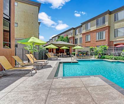 The gorgeous, large, turquoise outdoor pool with pool chairs, tables, and chairs with the tan and rust-colored apartments in the background at Visions Apartment Homes. 