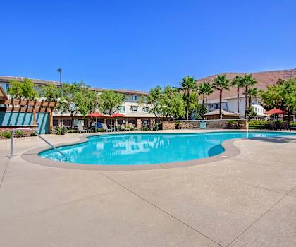 The large, turquoise outdoor pool surrounded by palm trees, pool chairs, tables, cabanas, and chairs with the apartments in the background at Vesada Apartment Homes. 