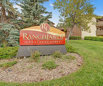 The exterior sign with trees and surrounded by greenery with the buildings in the background of Ranchlands Apartment Homes. 