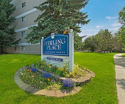 The blue exterior sign with trees and surrounded by greenery and flowers with the buildings in the background of Stirling Place Apartment Homes. 