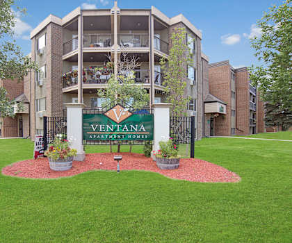 The emerald green exterior sign with trees and surrounded by greenery with potted flowers in front and the buildings in the background of Ventana Apartment Homes. 