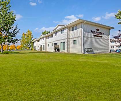 The beautiful exterior of the white apartments with a sign on the building and the green space with grass, trees, and shrubs at Westhaven Estates Apartment Homes. 