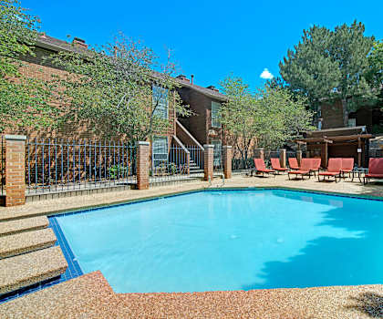 The large, turquoise outdoor pool with pool chairs, tables, and chairs with the apartments in the background at Windtree Apartment Homes. 
