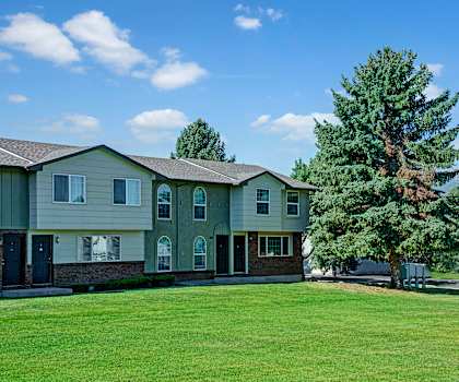 The exterior of the apartments and the green space with grass, trees, and shrubs and mountains in the background at Yorkshire Square Apartment Homes. 