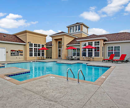 The large, turquoise outdoor pool with pool chairs, tables, and chairs with the apartments in the background at the Club at Heritage Apartment Homes. 