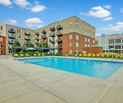 The large, turquoise outdoor pool with lime green pool chairs, tables, and blue chairs with the apartments and trees in the background at the Covington Apartment Homes.