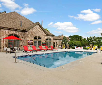 The large, turquoise outdoor pool with red pool chairs, tables, and chairs with red umbrellas with the apartments in the background at the Fairways at Edinburgh Apartment Homes. 