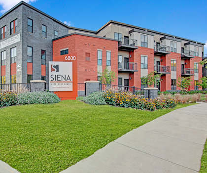 The exterior of the orange and gray Siena Apartment Homes with a white sign and grassy area with flower beds in front. 