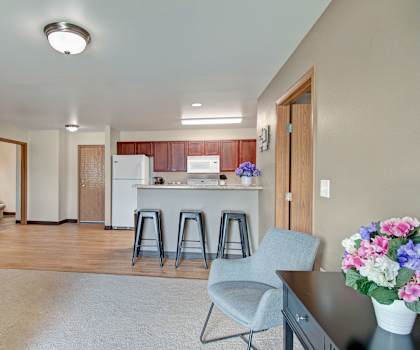 The living room with a couch, plush chairs, coffee table, decorations, a TV, and view of the kitchen at Pheasant Ridge Apartments.