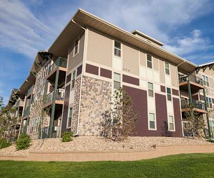 The exterior of the apartments and the green space with grass, trees, and shrubs at Prairie Pines at the Ridge Apartment Homes. 