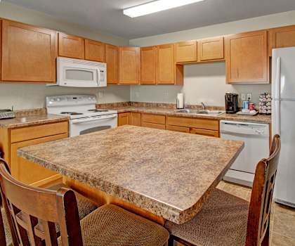 A spacious kitchen with white appliances, kitchen utensils, overhead lighting, light countertops with lots of room, a bar counter with barstools, and large wooden cabinets at Roosevelt East Apartment Homes. 